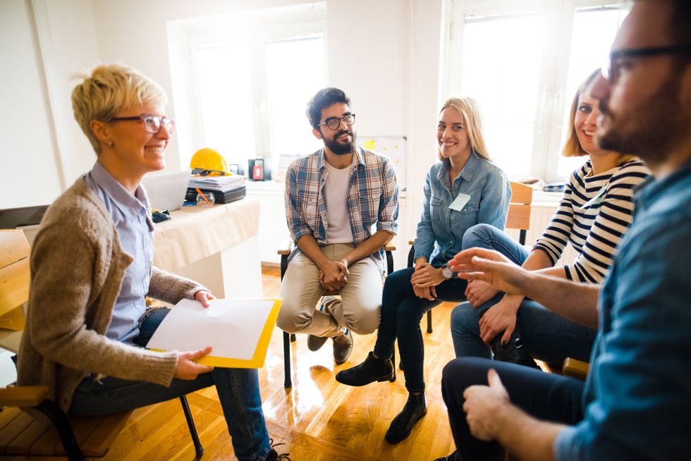 Two men and women sit in a semi-circle facing a woman with blond short hair. They're all listening and smiling.