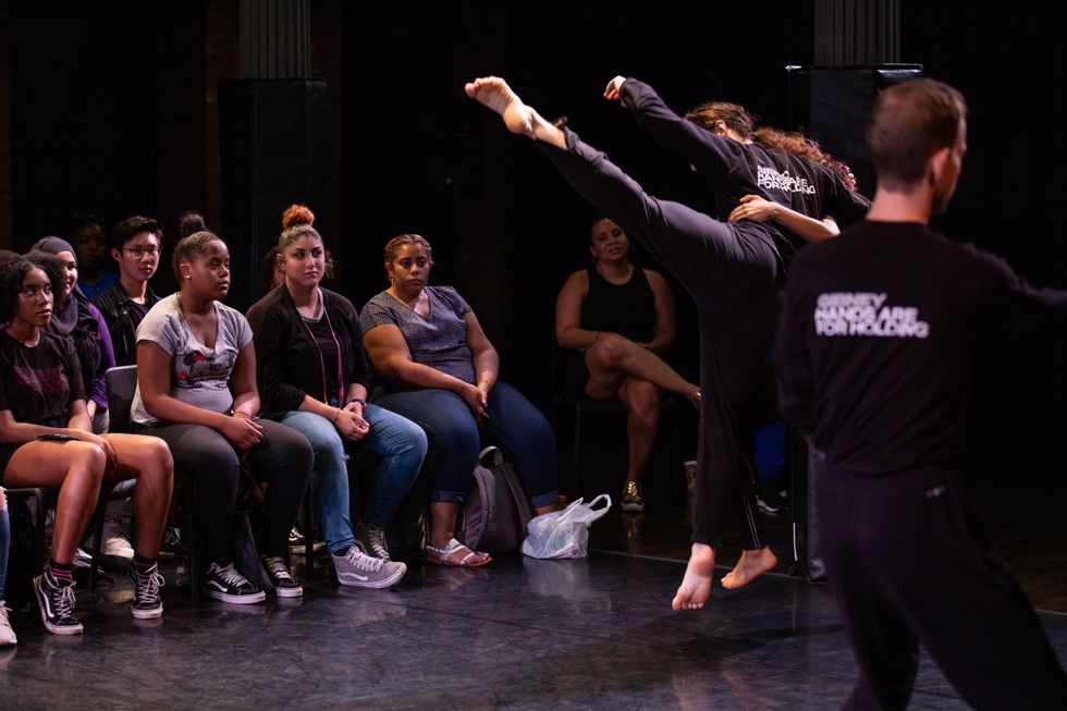 A view from the stage. As modern dancers perform, a seated audience of young adults looks on.