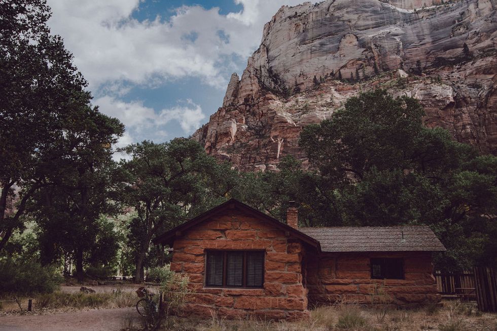 A rustic 1925 stone cabin, nestled around scrub and trees. A rock formation towers in the background.