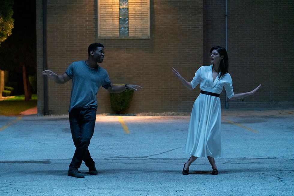 A young man and woman dancing in an empty parking lot. They each have one leg crossed in front of the other, with their hands out to their sides. They look at each other and smile. She wears a long white belted dress, he wears jeans and a grey t shirt.