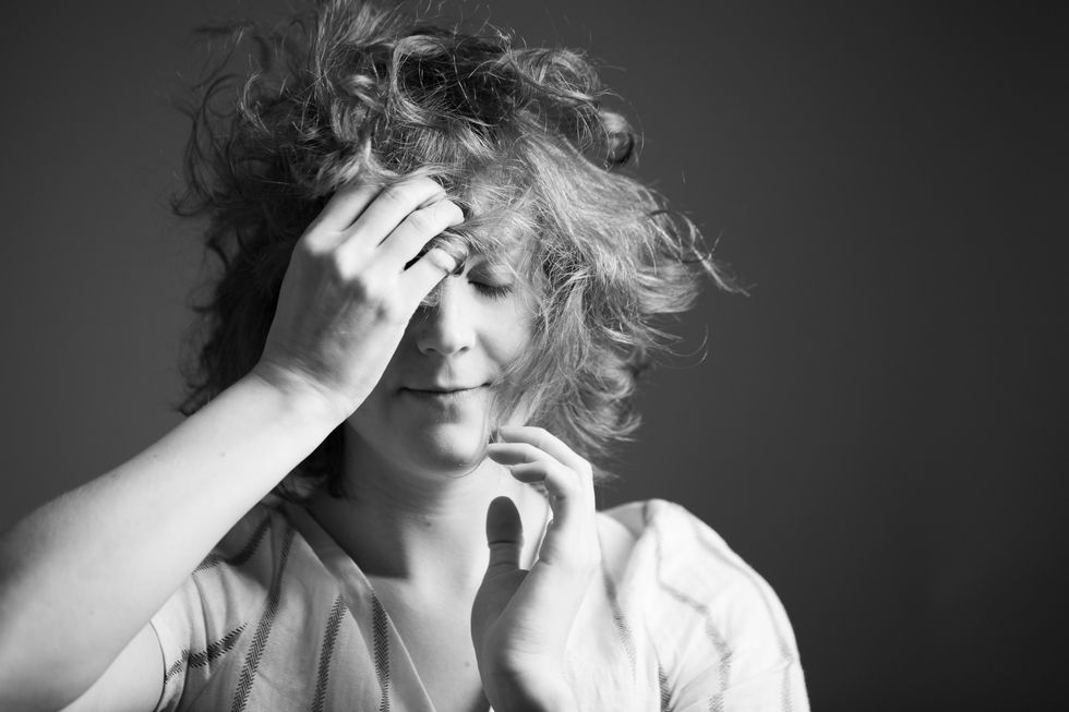 In a black and white image, Ladenheim, hair curling wildly around her face, closes her eyes and smiles gently as she brings the fingertips of one hand to her forehead, the others to her chin.