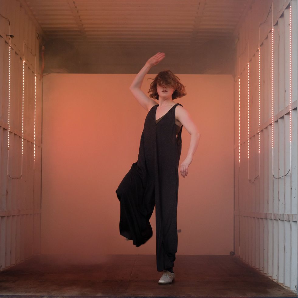 A woman in a draping black jumpsuit stands on one leg, the same arm raised alongside her head, chin-length hair flying into her eyes.
