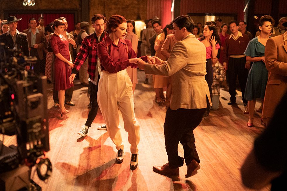 Tommy Tonge, dressed in a flannel shirt and Converse sneakers, walks around Natalie Dormer and Johnathan Nieves, both dressed in period-appropriate suits, as they clasp hands mid-swing. A camera is visible to the left, and extras in similar attire look on.