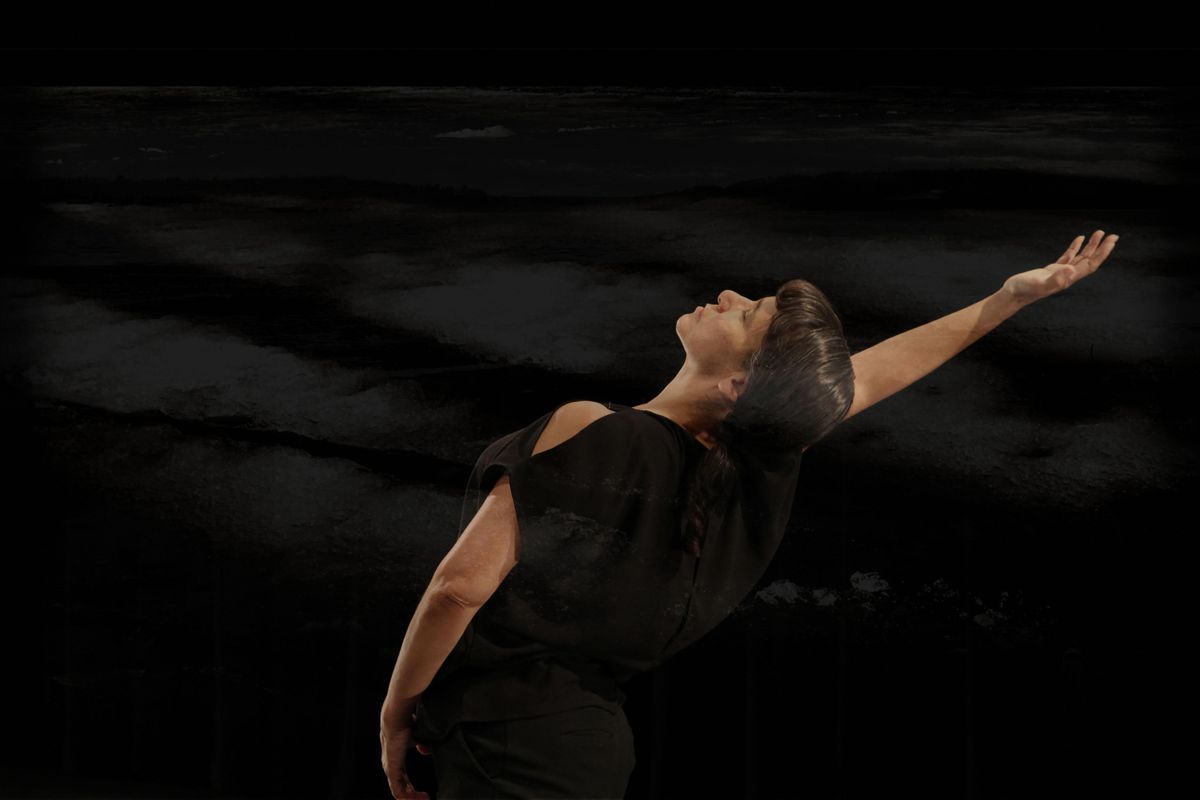 Simas wears a black shirt and pants and stands against a black background. Her body is tilted backwards with her right arm extended behind her. She has dark hair and her eyes are closed.