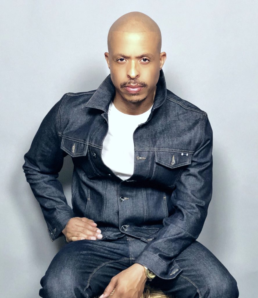 Jamal Smith, in a white t-shirt and denim jacket, gazes at the camera in front of a grey background.