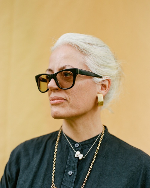 A headshot of Castro, who looks off serenely toward the left of the frame. Her white hair is pulled back; she wears a black button-up shirt, gold jewelry, and smoky sunglasses.