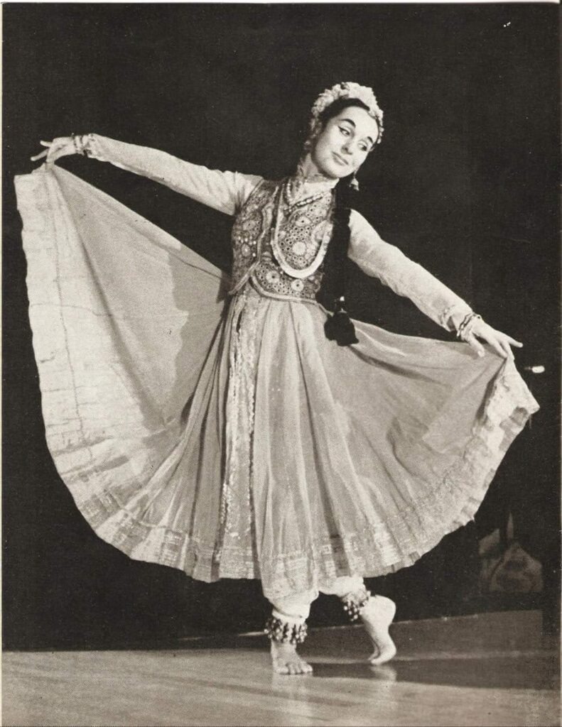 A sepia-toned photo of a young Vetra in classical Indian dress. She holds out the ends of her full skirt, looking over her right shoulder, her right foot tucked behind her left leg.