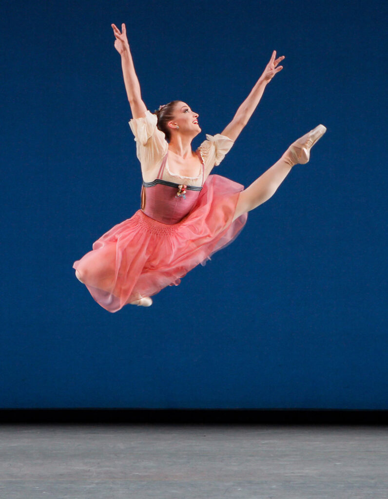 Bouder, wearing a candy-pink ballet dress with a long skirt and white blouson sleeves, is caught at the height of an enormous Italian pas de chat, with her left leg extended and both arms flying.