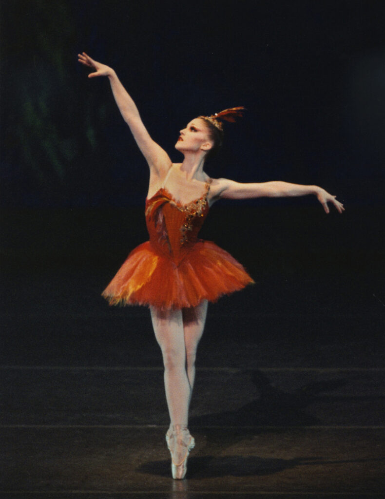 Bouder, wearing a feathered red tutu and headpiece, stands in sous-sus on pointe, looking up and out over her raised right arm.