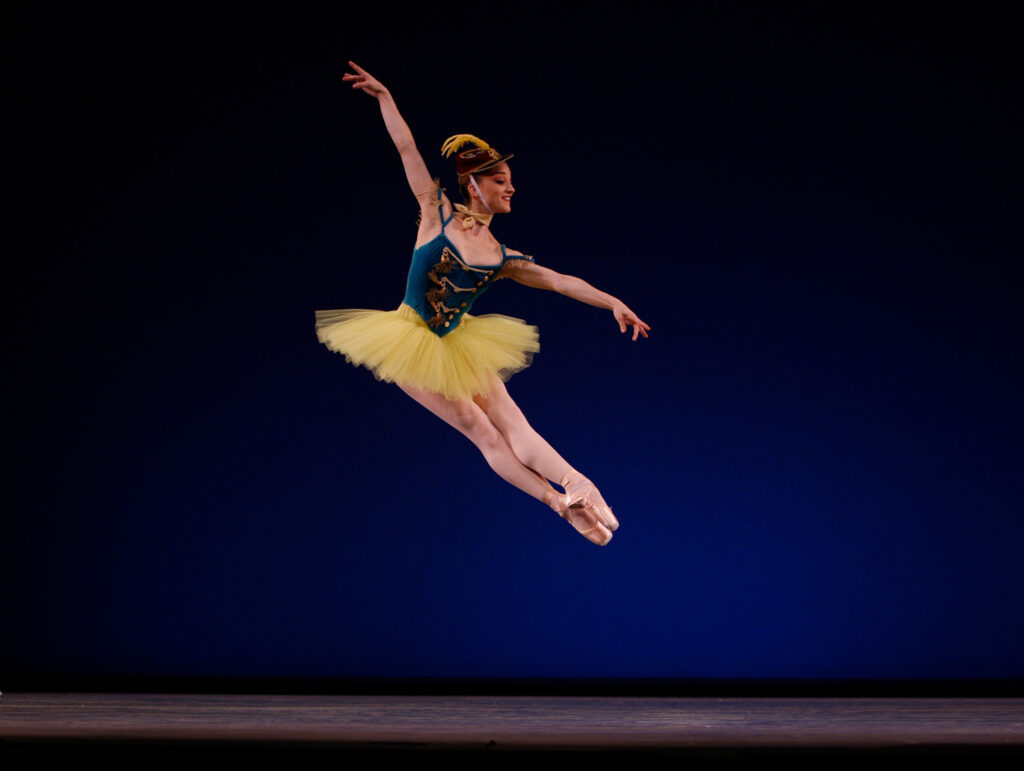Bouder, wearing a tutu with a blue velvet bodice and yellow skirt and a red military cap with a yellow feather, is pictured at the height of an assemblé, her body leaning forward over her outstretched legs.