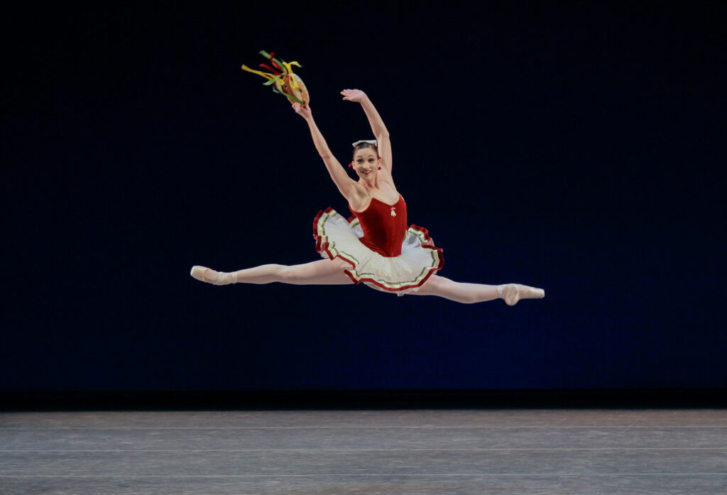 Bouder, wearing a tutu with a red velvet bodice and white ribbon-lined skirt, is caught at the height of a huge saut de chat, her face beaming. The ribbons on the tambourine that she holds in her right hand stream out behind her.