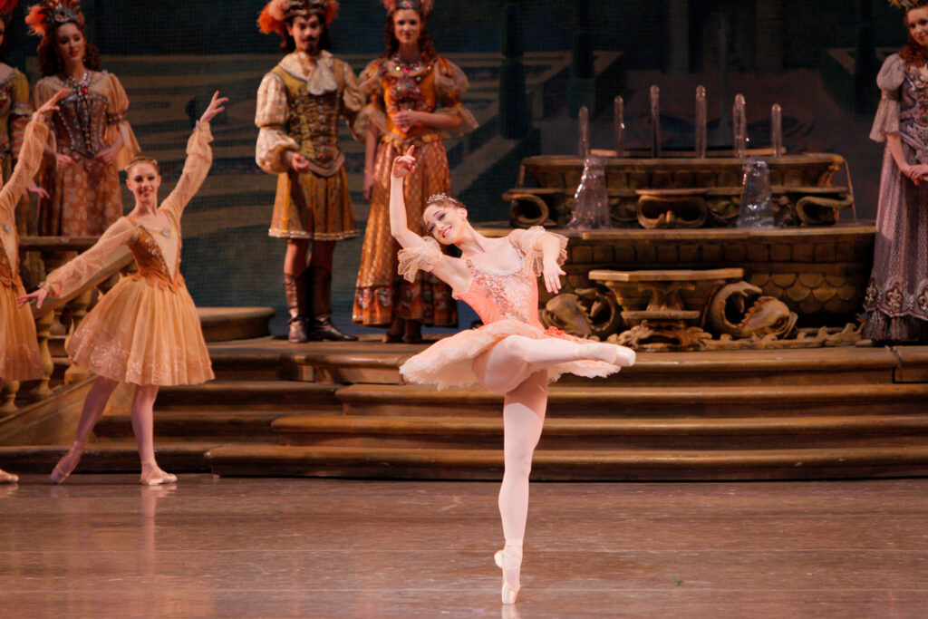 Bouder, wearing a pink tutu with flower details, stands on pointe in attitude devant, leaning back luxuriantly into her raised right arm.