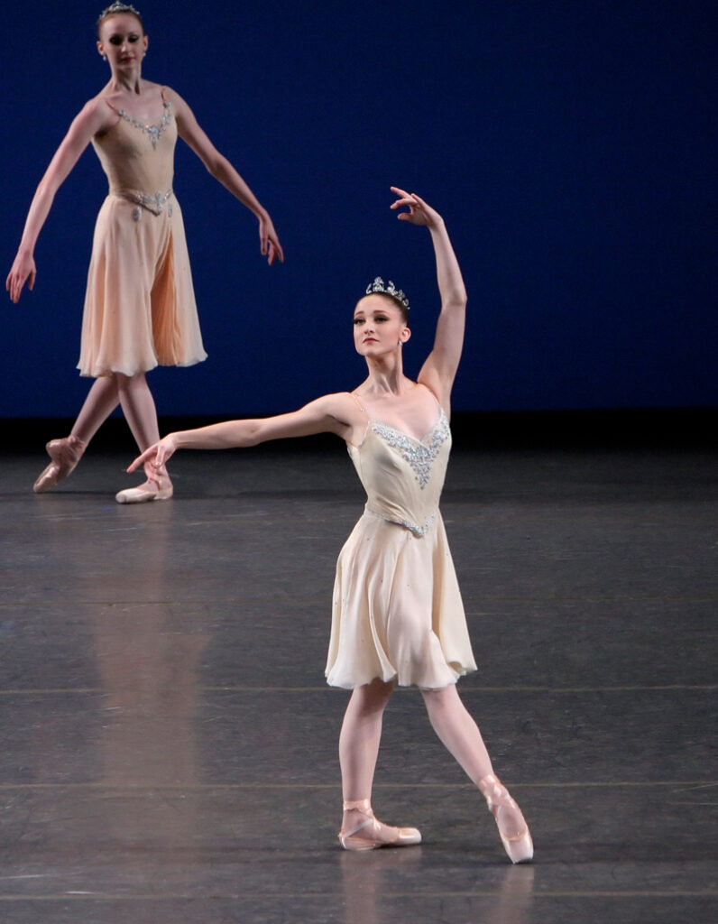 Bouder, wearing a flowing white dance dress and a tiara, stands in a majestic tendu devant.